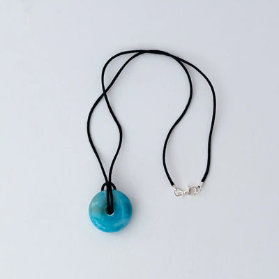 Pierre naturelle de larimar taillée en forme de donut montée sur un fil noir avec fermoir en argent 925. Larimar de couleur bleu, collier posé sur un fond blanc.